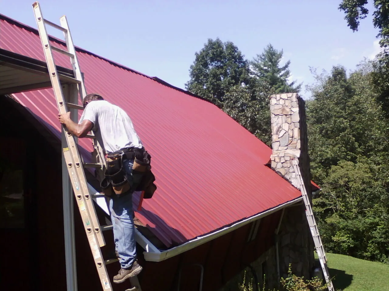 Roofer on a ladder beside a red metal roof and stone chimney — Soffit & Fascia Repair by Roofing Brothers in Chuckey